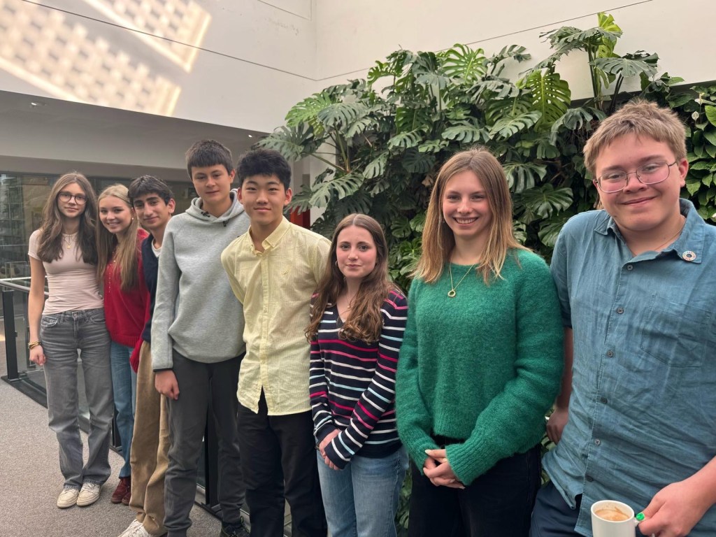 The internship cohort standing together indoors in front of a wall of green foliage