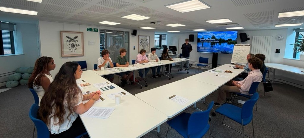 Participants seated around a large open square of tables in a meeting room, with a presentation screen reading Fenlands Restoration for Flood Defence and Water Quality and a flip chart headed Youth Workshop