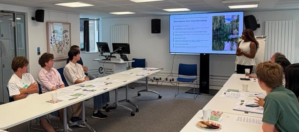 A young person presenting a slide titled Restoring Our Fens: School Workshops to participants seated around tables at the working session