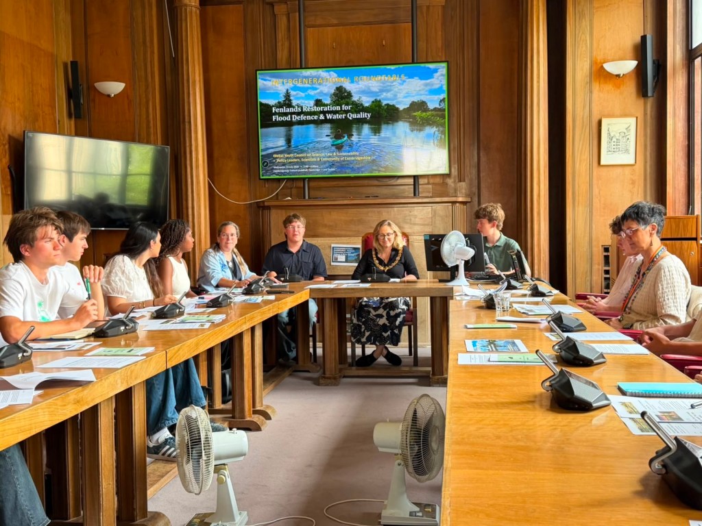 Young people and councillors seated around the wood-panelled council chamber table with microphones, beneath a screen reading Intergenerational Roundtable: Fenlands Restoration for Flood Defence and Water Quality