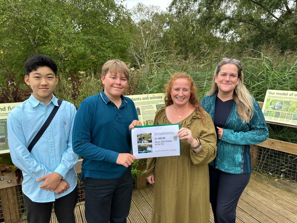 Four young people standing together beside an interpretation board on a boardwalk, holding printed materials