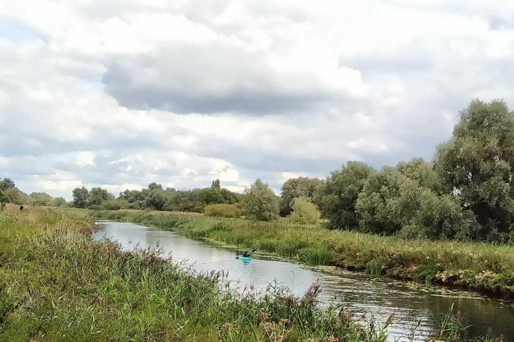 A narrow river winding through open fen meadow under a wide cloudy sky